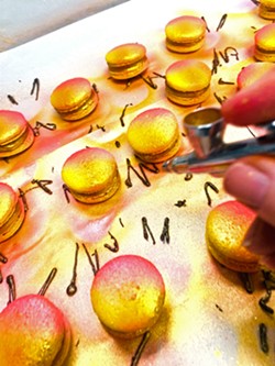 ARTIST AT WORK Monika Anderson's salted caramel macarons are airbrushed yellow, orange, and hot pink; dusted with 24-carat edible gold powder mixed with vodka; then topped off with pure dark chocolate and a pinch of salt crystals. - PHOTO COURTESY OF MONIKA'S MACARONS