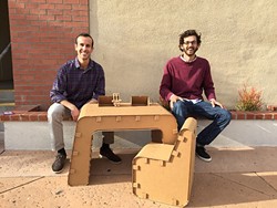 JUST ONE OF THE GUYS:  Jake Disraeli (left) and Justin Farr (right), owners of The Cardboard Guys, sit with their company&rsquo;s cardboard desk and chair with their logo and mascot, Cardboard Corey. - PHOTO BY REBECCA LUCAS